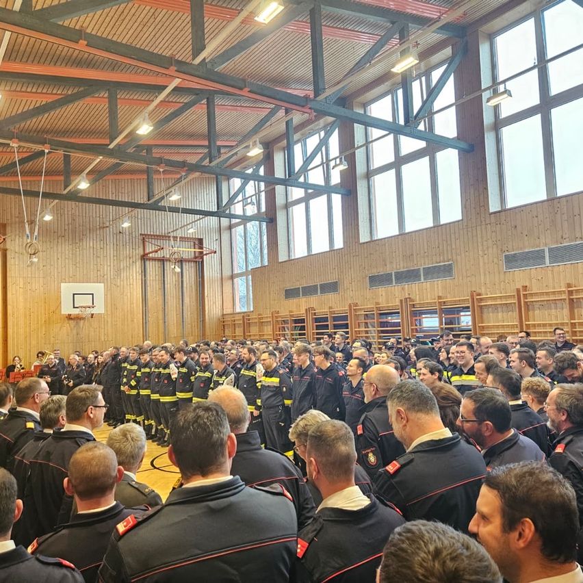 Eine große Gruppe von Feuerwehrleuten in Uniform steht in einer Turnhalle, die auf eine Bühne mit einer Frau blickt, die Trompete spielt. Die Halle hat Holzwände, einen Basketballkorb und helle Deckenleuchten.
