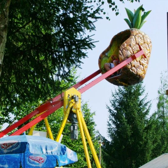 Eine Person fährt auf einer rot-gelben Achterbahn mit Ananas-Thematik, unter einem blauen Schirm mit dem Corona-Logo.
