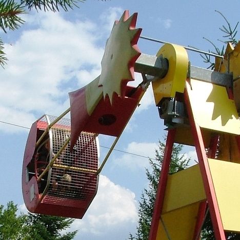 Ein farbenfroher Vergnügungspark, der eine rot-gelbe Struktur mit einer schwingenden Gondel zeigt, umgeben von üppigen grünen Bäumen und blühenden Blumen unter einem blauen Himmel.