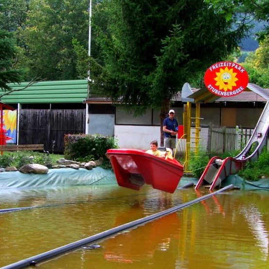Eine rote Bootsfahrt im Freizeitpark Stubenbergsee ist teilweise im Wasser versunken, mit zwei Kindern im Inneren und einem Mann in der Nähe.