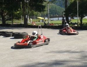 Zwei Personen rasen in roten Go-Karts auf einer asphaltierten Strecke, mit Bäumen und einem See im Hintergrund. Sie tragen weiße Helme.
