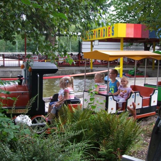 Vier junge Kinder fahren mit einem Miniaturzug mit gelbem Dach in einem Park. Bäume und ein buntes Gebäude sind im Hintergrund zu sehen.