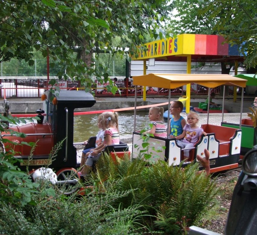 Vier junge Kinder fahren mit einem Miniaturzug mit gelbem Dach in einem Park. Bäume und ein buntes Gebäude sind im Hintergrund zu sehen.