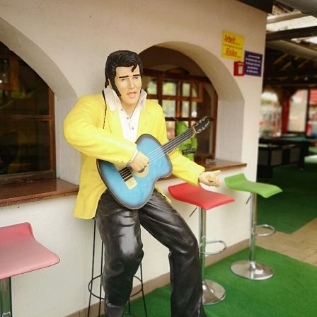 Eine Statue von Elvis Presley, die auf einem Hocker sitzt und eine blaue Gitarre hält, vor einer Theke. Drei bunte Hocker stehen dahinter.