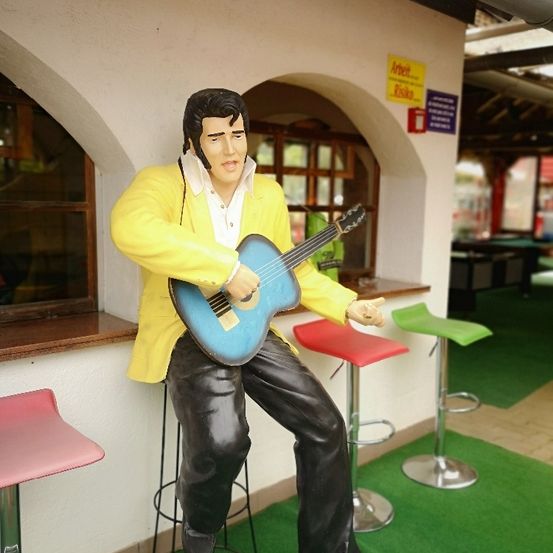 Eine Statue von Elvis Presley, die auf einem Hocker sitzt und eine blaue Gitarre hält, vor einer Theke. Drei bunte Hocker stehen dahinter.