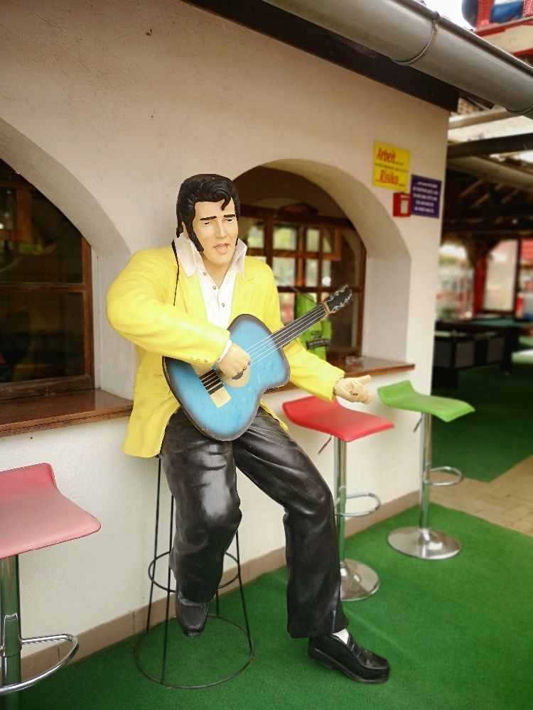 Eine Statue von Elvis Presley, die auf einem Hocker sitzt und eine blaue Gitarre hält, vor einer Theke. Drei bunte Hocker stehen dahinter.