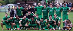 Fußballmannschaft in grünen Trikots feiert auf dem Feld, mit Trainern und Fans hinter ihnen, unter einem grünen Baldachin.