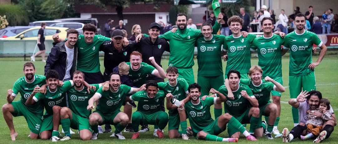 Fußballmannschaft in grünen Trikots feiert auf dem Feld, mit Trainern und Fans hinter ihnen, unter einem grünen Baldachin.