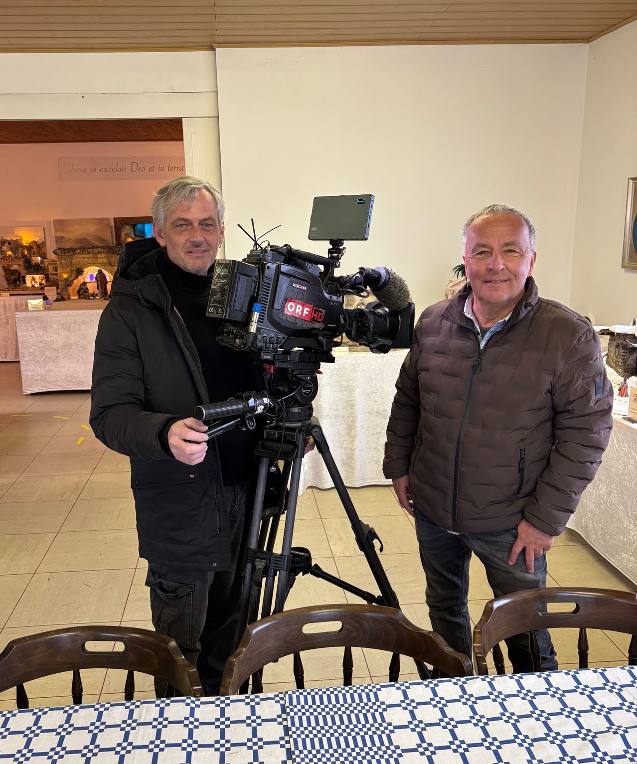 Zwei Männer stehen neben einer Videokamera mit Stativ in einem Raum. Ein Mann lächelt und hält das Stativ. Hinter ihnen befindet sich eine weiße Wand mit einem Bilderrahmen. Es gibt Stühle und einen Tisch mit einer weißen Tischdecke.