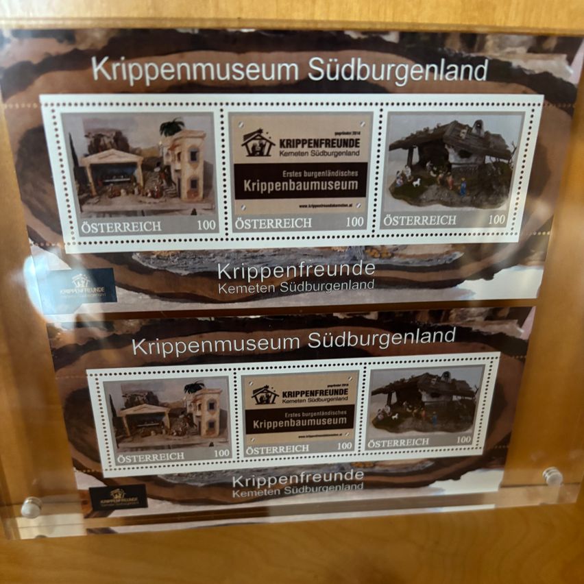 Ausstellung von zwei Sätzen von Briefmarken aus dem Krippenmuseum Südburgenland. Jeder Satz enthält drei Briefmarken, eine mit einem Gebäude, eine mit Text und eine mit einer Weihnachtskrippe. Die Briefmarken sind aus Österreich und haben einen Wert von 100.