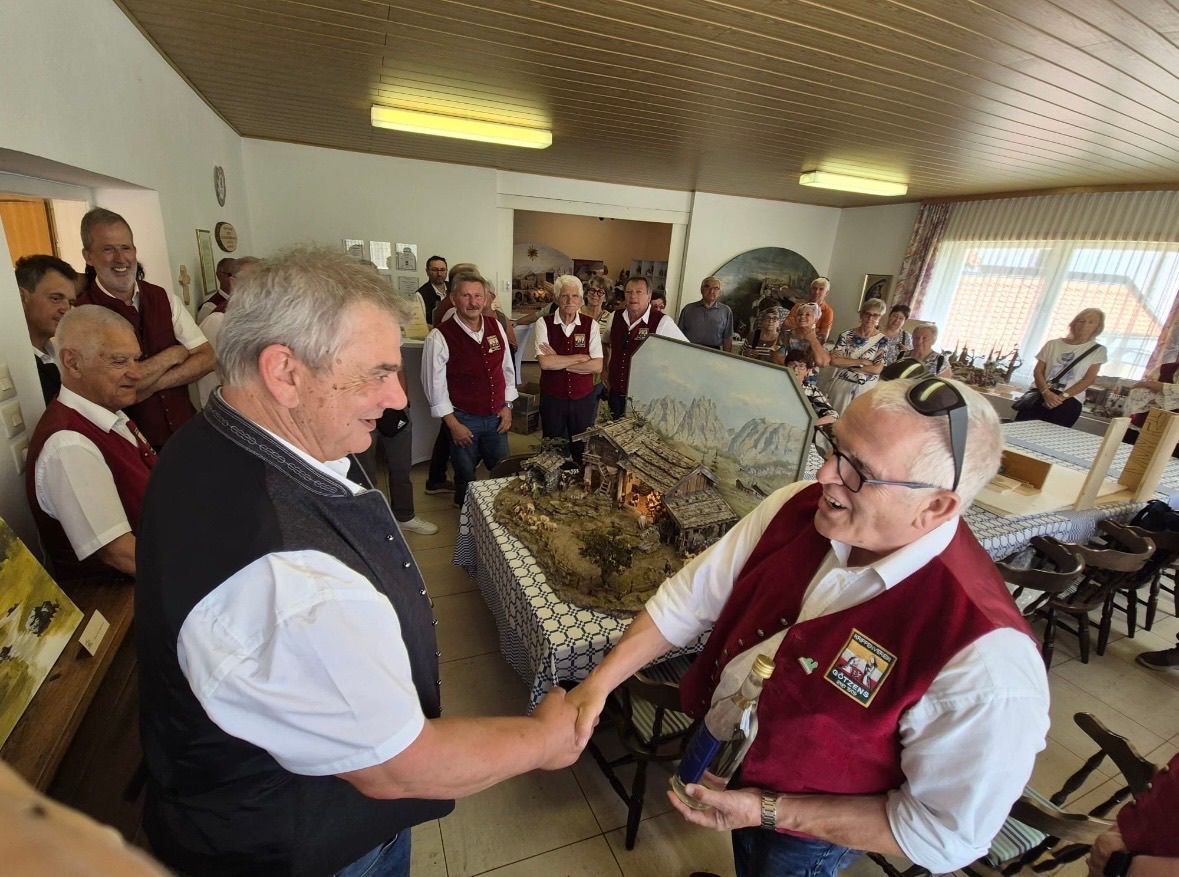 A group of people are gathered around a table in a room. Two men are shaking hands. The man on the right is holding a bottle. There is a table with a cake on it and a frame on the wall.