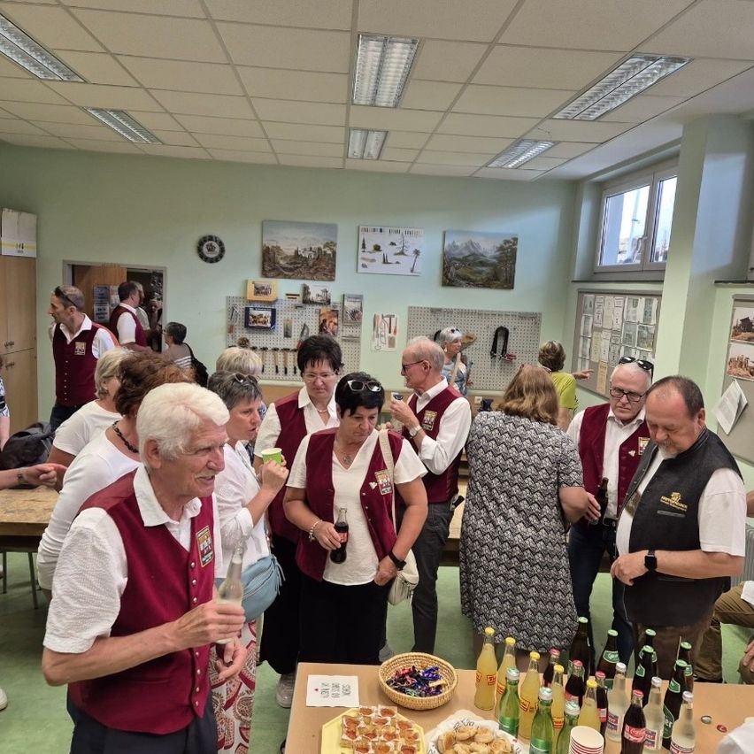 A group of adults in red vests stand in a room. Some are holding drinks. A table with snacks and drinks is in the foreground. Pictures and a clock are on the wall.