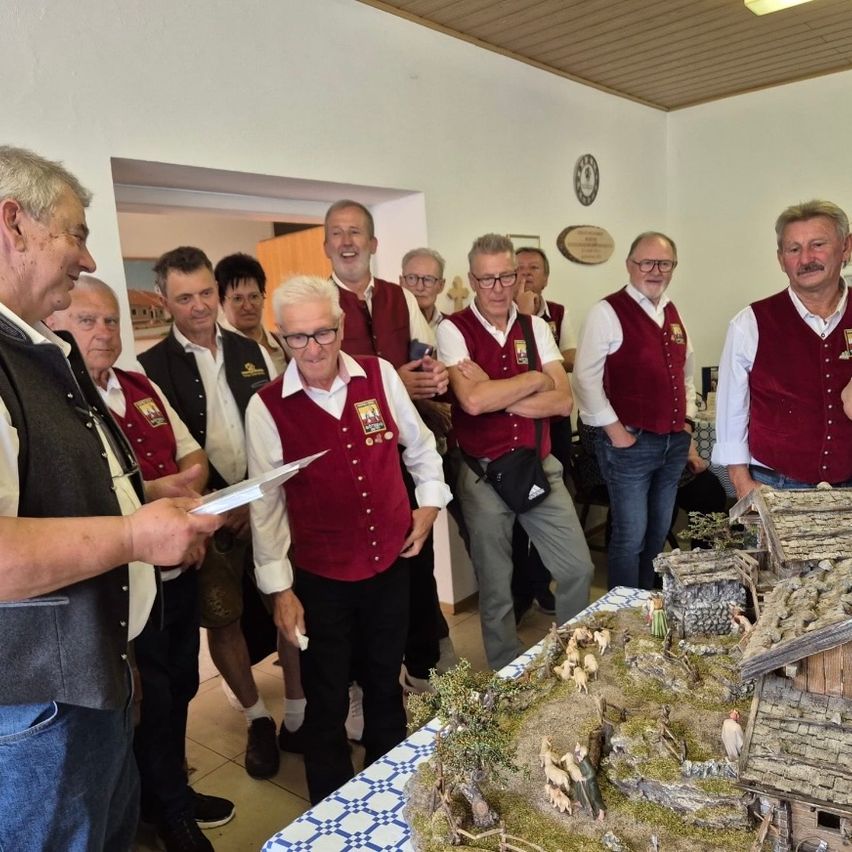 A group of men wearing red vests are standing in a room with a man reading a piece of paper. The room has a wooden ceiling and a tiled floor. There is a wooden structure with a roof on a table.