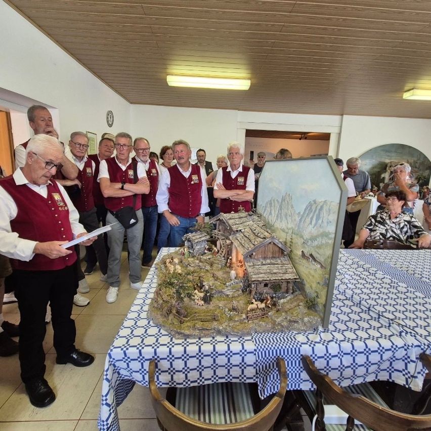 A group of older men in vests stands around a table with a model village scene on it. A man holds papers and reads aloud.