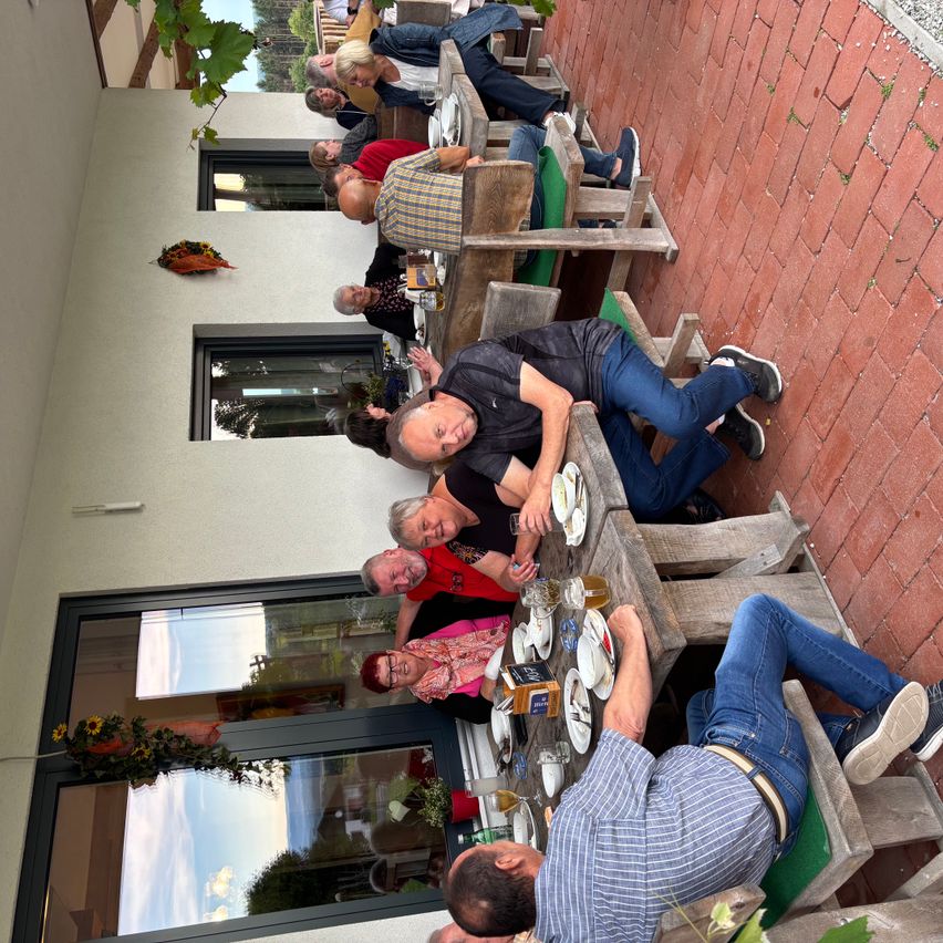 Eine Gruppe von Menschen sitzt um einen Tisch auf einer Terrasse mit Essen und Getränken. Einige Leute tragen Turnschuhe.