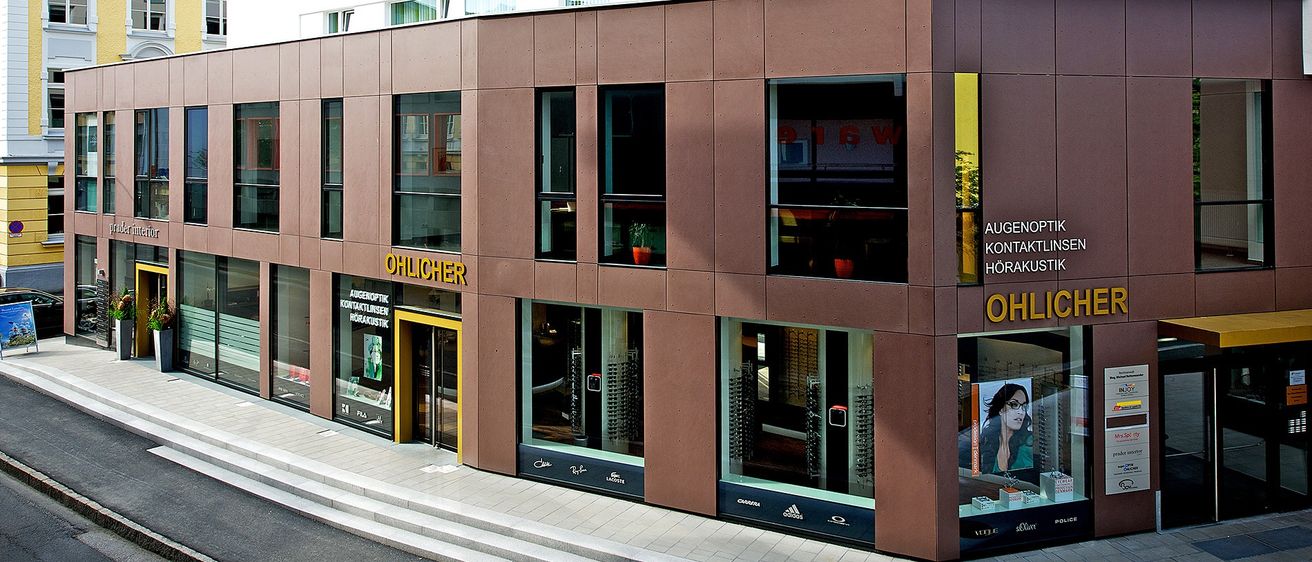 Ein braunes Ladenlokal mit dem Namen 'Ohlicher' darauf. Der Laden hat große Glasfenster. Ein Bild einer Frau mit Brille ist im Schaufenster zu sehen.