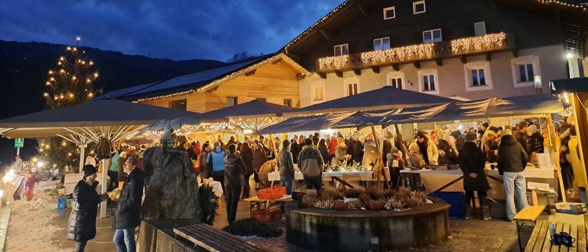 Ein geschäftiger Weihnachtsmarkt bei Nacht unter einem bewölkten Himmel. Viele Menschen kaufen an Ständen mit großen Regenschirmen ein. Ein geschmücktes Haus und ein Weihnachtsbaum sind im Hintergrund zu sehen.