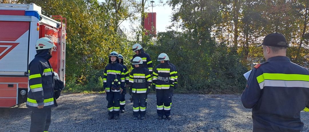 Eine Gruppe junger Menschen in Feuerwehruniformen steht im Kreis auf einer Kiesfläche. Sie tragen Helme und werden wahrscheinlich ausgebildet. Ein Feuerwehrausbilder steht in der Nähe.