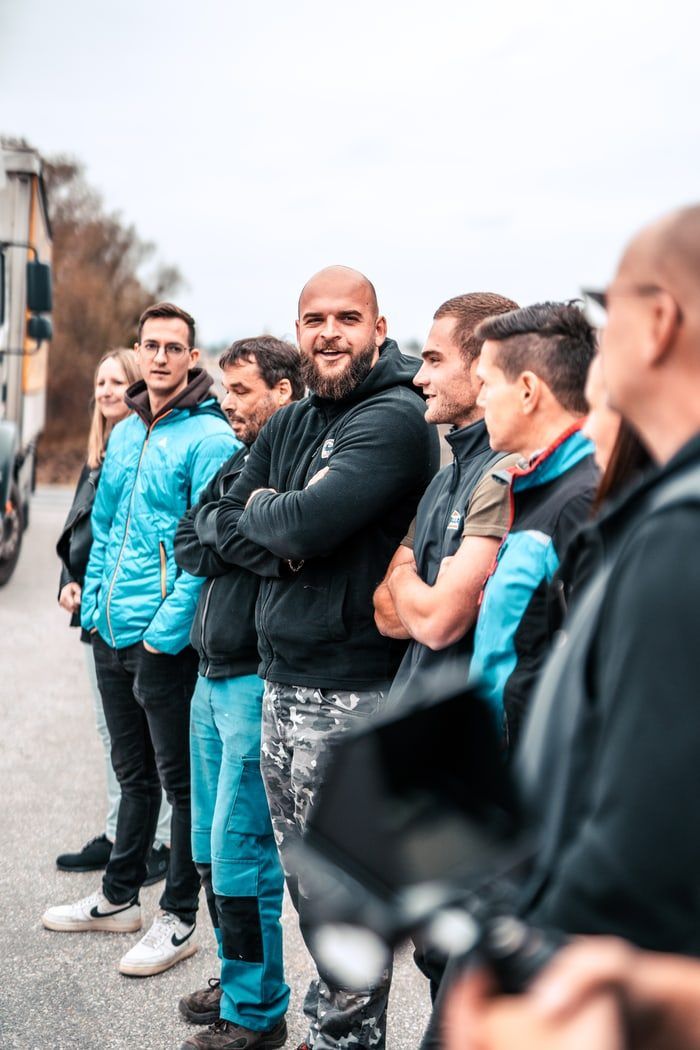 Eine Gruppe von Menschen in blauen und schwarzen Jacken steht draußen zusammen, mit einem Bus im Hintergrund. Sie scheinen in Gespräche vertieft zu sein.