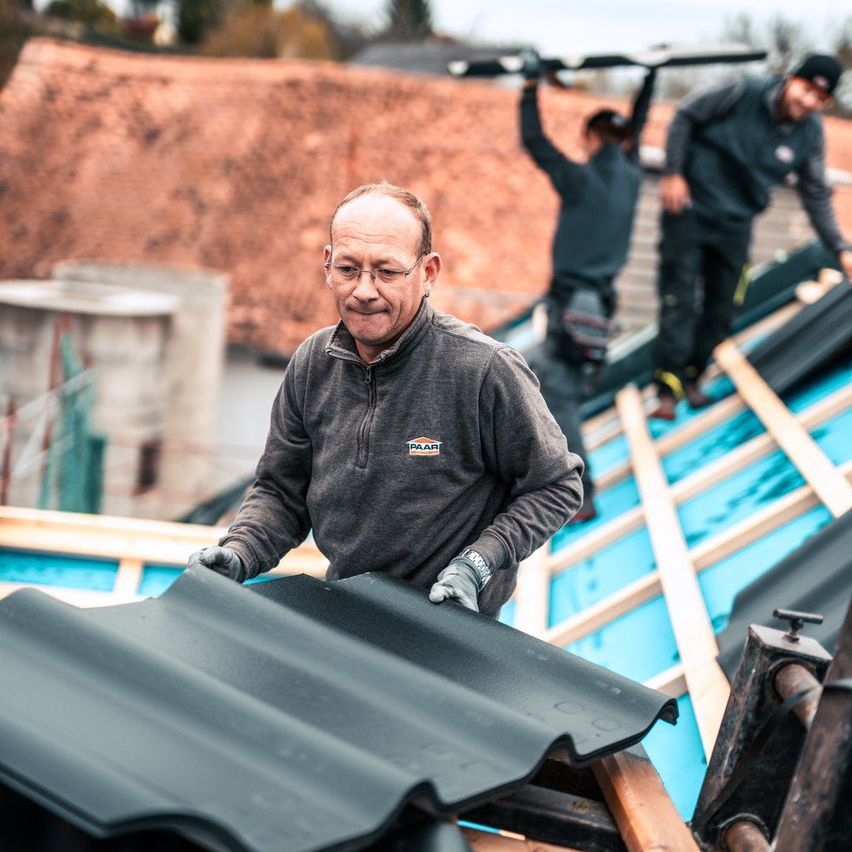 Ein älterer Mann mit Brille installiert eine schwarze Dachplatte auf einer Baustelle. Zwei weitere Arbeiter stehen auf dem Dach.