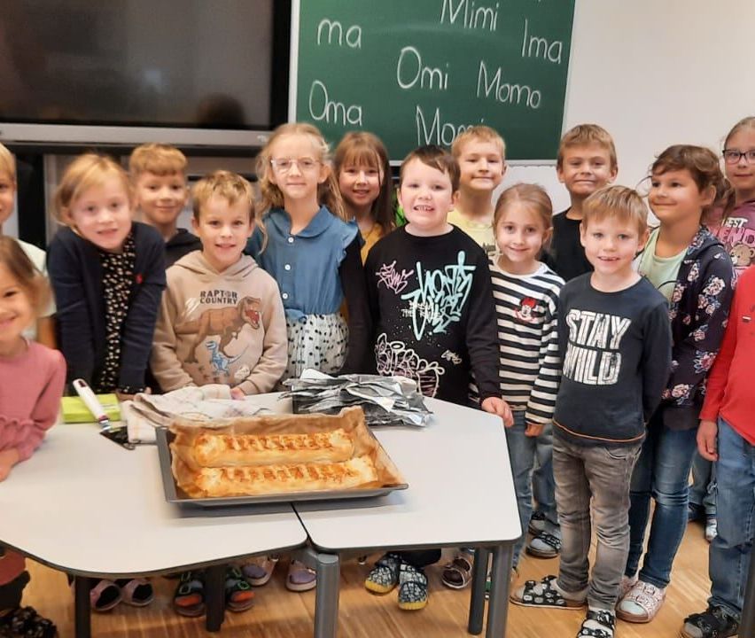 Eine Gruppe von Kindern steht in einem Klassenzimmer mit einer Tafel dahinter. Ein Tisch hat ein Lebensmittel darauf, und die Hand einer Person ist teilweise auf der linken Seite zu sehen.