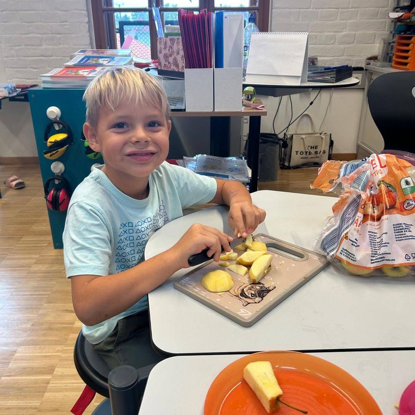 Ein junger Junge mit blonden Haaren lächelt, während er Äpfel auf einem Tablett schneidet. Er sitzt an einem Schreibtisch im Klassenzimmer.