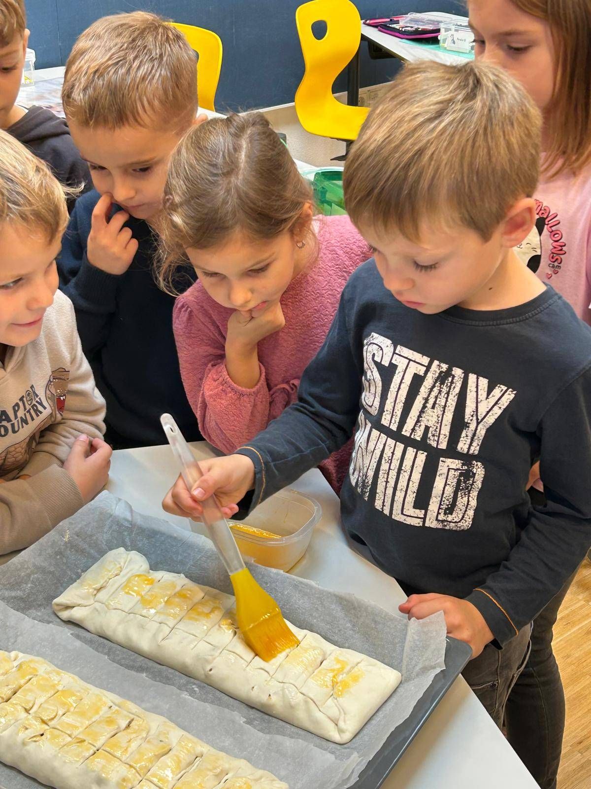 Eine Gruppe von Kindern ist in einer Backaktivität beschäftigt. Sie bestreichen ein Gebäck mit einer gelben Substanz auf einem Backblech. Ein Kind trägt ein blaues Shirt mit weißem Text.
