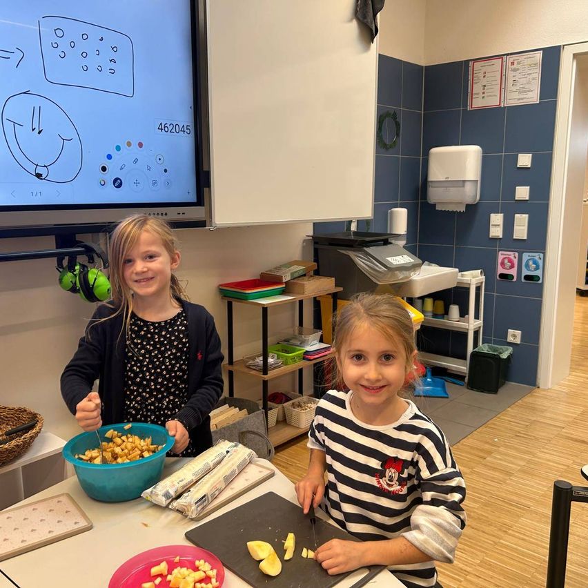 Zwei Mädchen sind in einem Klassenzimmer. Eine mischt Essen in einer Schüssel und die andere schneidet Essen auf einem Schneidebrett. Ein Monitor ist an der Wand mit einer Zeichnung.