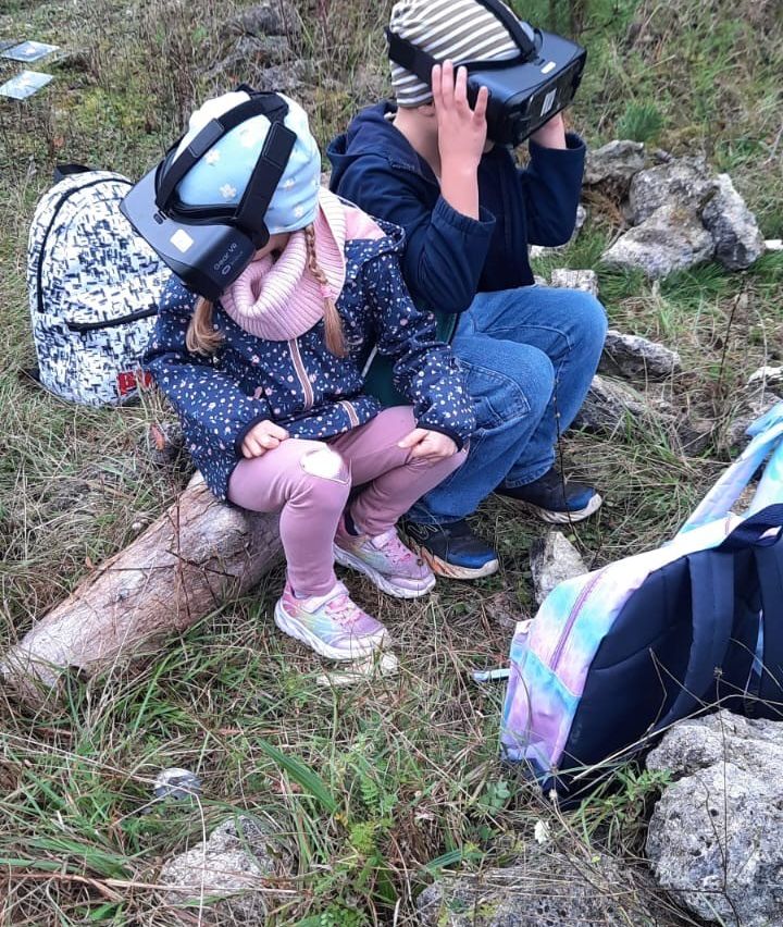 Zwei Kinder benutzen VR-Headsets. Eines sitzt auf einem Baumstamm, das andere neben ihnen. Sie sind von Rucksäcken, Gras und Steinen umgeben.