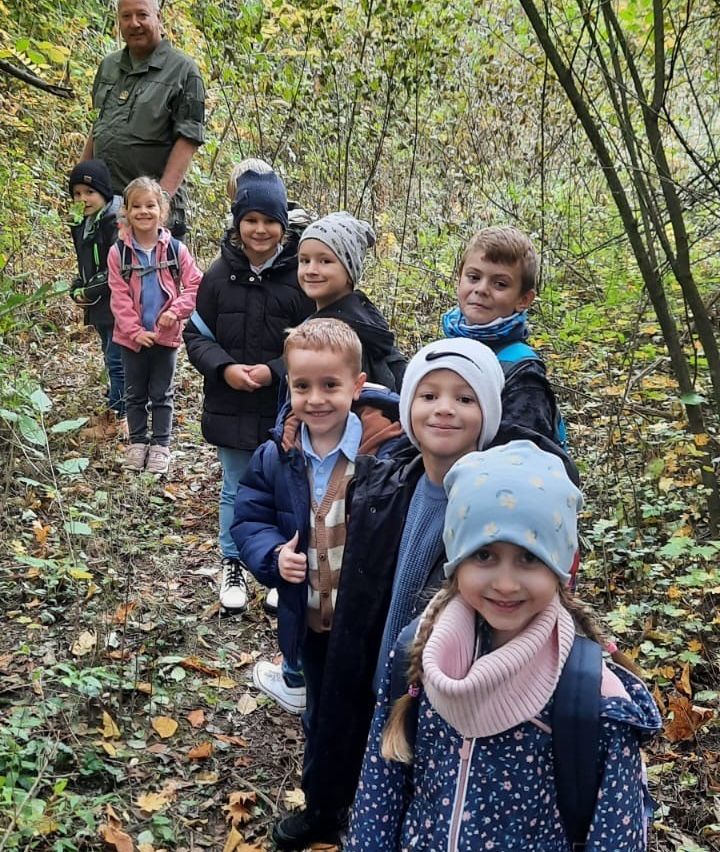 Eine Gruppe von Kindern und einem Erwachsenen wandert tagsüber in einem bewaldeten Gebiet. Die Kinder lächeln und scheinen die Erfahrung zu genießen.