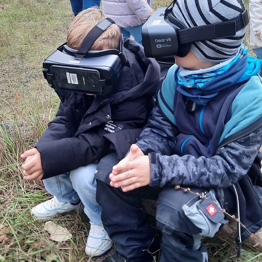 Zwei kleine Kinder sitzen auf dem Gras und tragen VR-Headsets. Sie tragen Winterkleidung und eines hat eine Schweizer Flagge auf seiner Jacke.