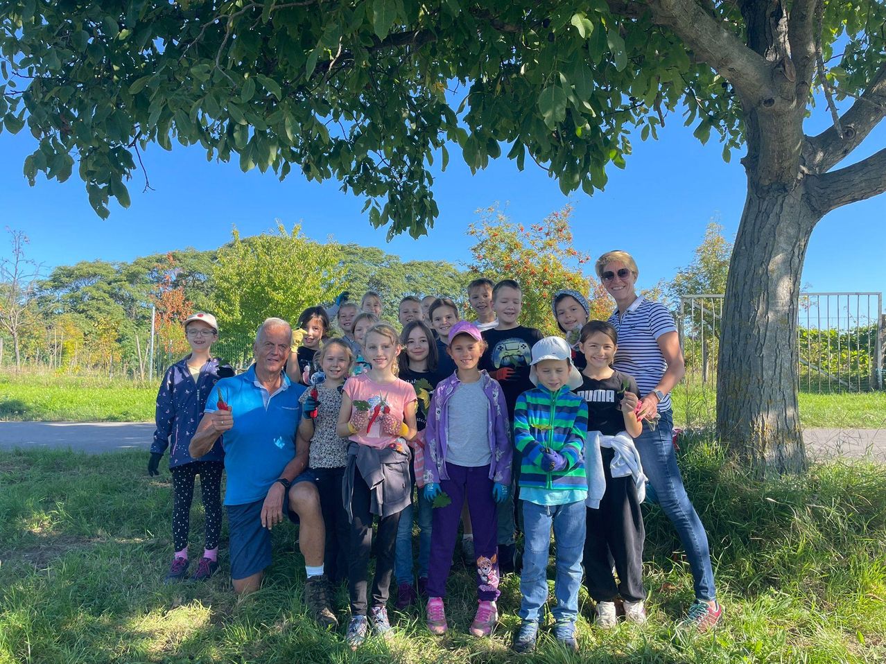 Eine Gruppe von Kindern und Erwachsenen posiert für ein Foto in einem Park. Sie stehen unter einem Baum, einige halten Pflanzen. Die Erwachsenen tragen Hüte und Brillen.