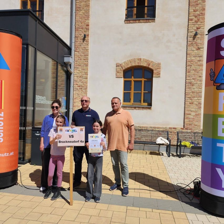 Fünf Personen stehen vor einem Gebäude mit Ziegelwänden. Zwei Mädchen halten Urkunden. Ein Mann trägt eine Sonnenbrille, ein anderer ein Polohemd. Zwei Frauen tragen Jacken.