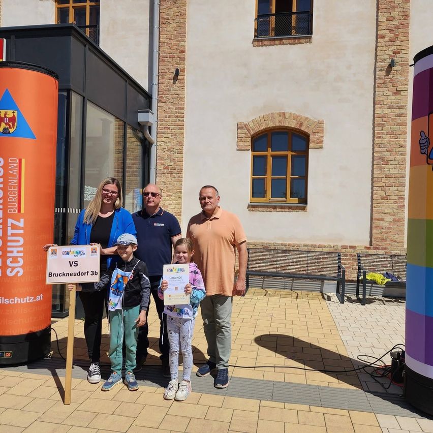 Vier Personen stehen vor einem Gebäude mit orangefarbenen Bannern. Zwei Kinder halten Urkunden in den Händen, und zwei Erwachsene stehen neben ihnen. Ein Gebäude mit Ziegelsteinen und Fenstern steht dahinter.