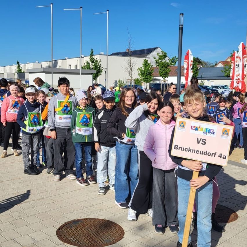 Eine Gruppe von Kindern in Uniform steht draußen, lächelt und posiert für ein Foto. Einige halten Schilder, und ein Junge vorne trägt einen Abzeichen. Gebäude und Autos sind im Hintergrund.