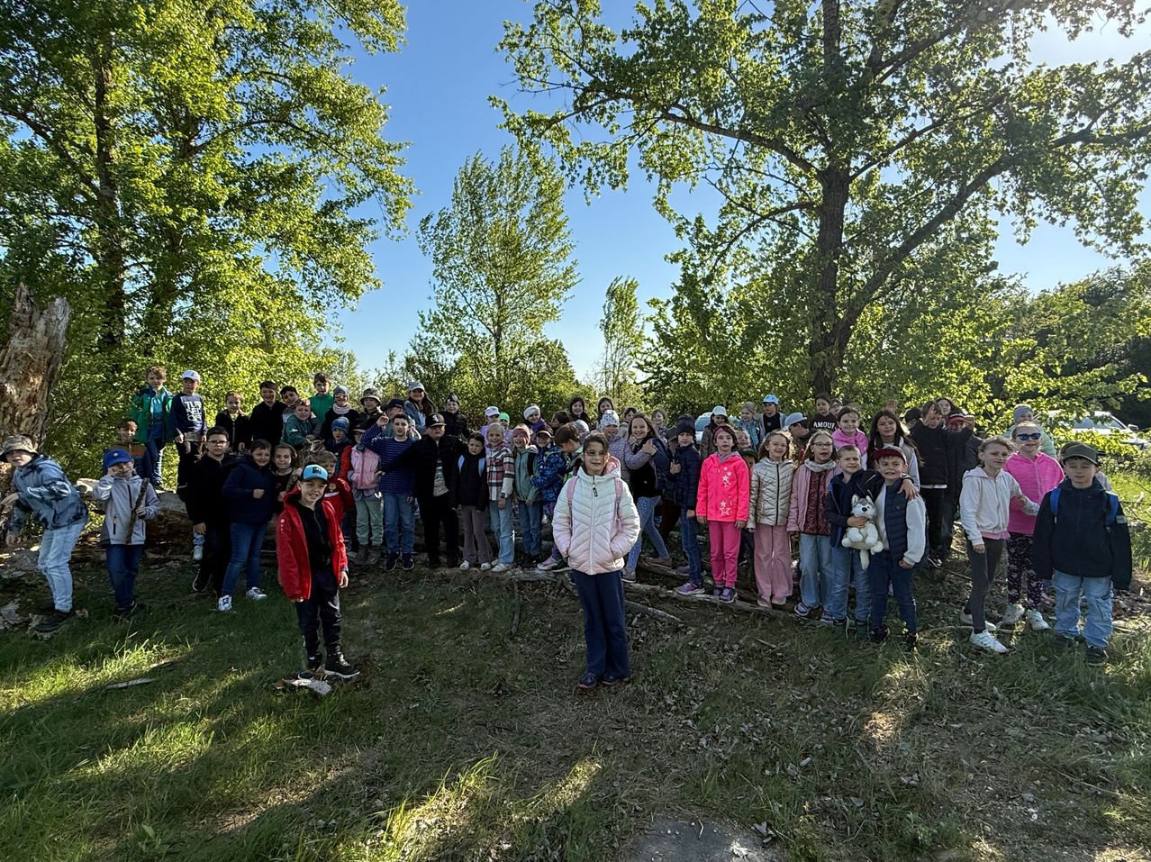 Eine große Gruppe von Kindern und Erwachsenen steht an einem sonnigen Tag im Freien, einige tragen Jacken, Mützen und Turnschuhe, mit Bäumen im Hintergrund.