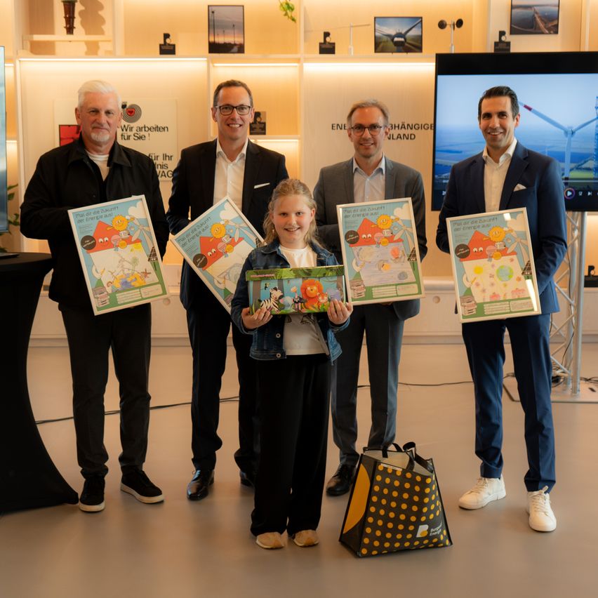Fünf Männer und ein Mädchen halten bunte Poster und lächeln, während sie gemeinsam posieren. Vor ihnen steht eine Tasche. Dahinter ist ein Fernseher eingeschaltet und zeigt eine Windmühle.