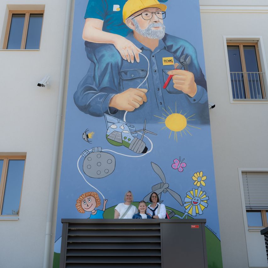 Ein Wandbild zeigt einen älteren Mann, der ein Kabel und ein Werkzeug hält. Darunter stehen zwei Frauen und ein Mädchen vor dem Wandbild. Das Gebäude hat mehrere Fenster und einen Balkon.