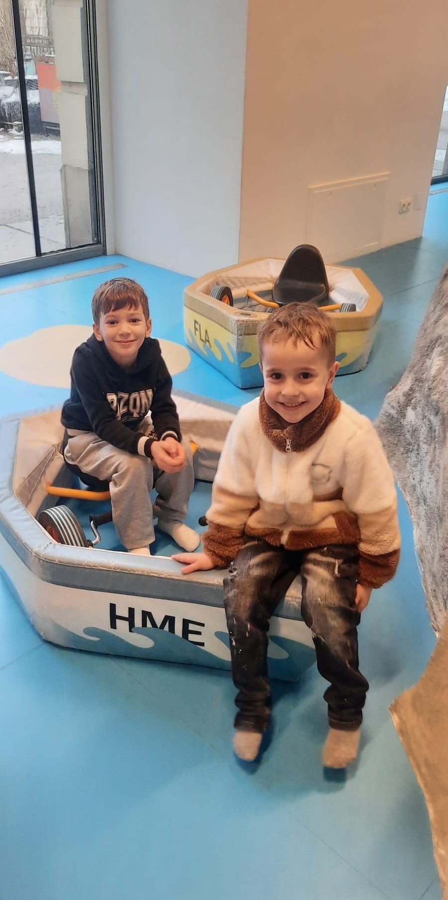 Two young boys are sitting in play boats, one with a black hoodie and the other with a white jacket. They are smiling and posing for a photo.