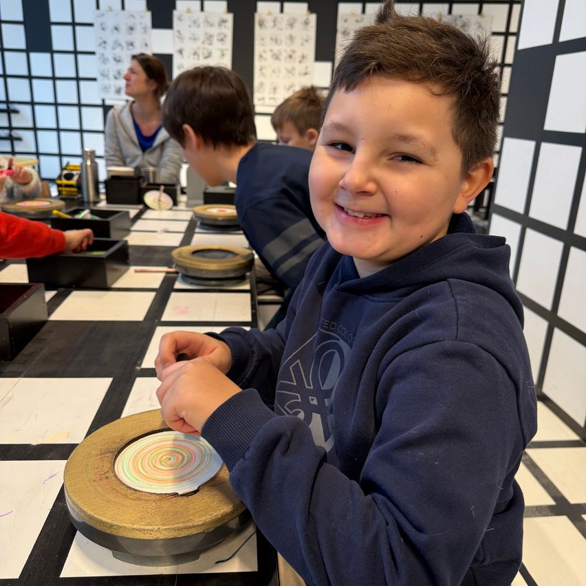A young boy smiles while holding a spinning top in a classroom with checkered floor and white walls.