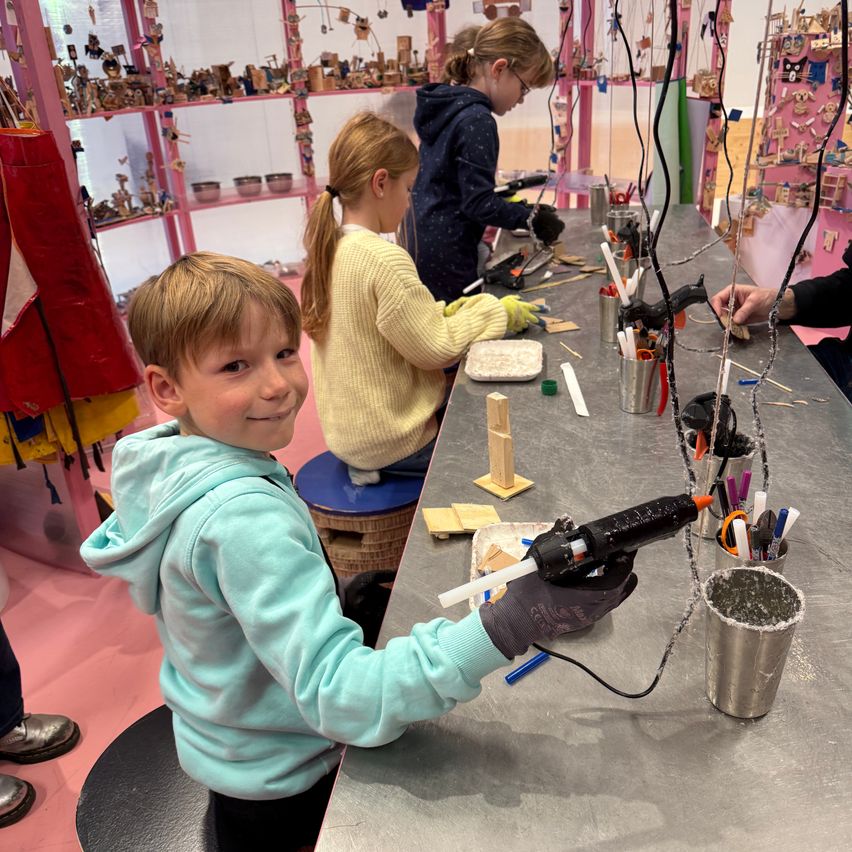 Children are working on a craft project in a workshop with various materials and tools. A boy in a blue hoodie is holding a hot glue gun.