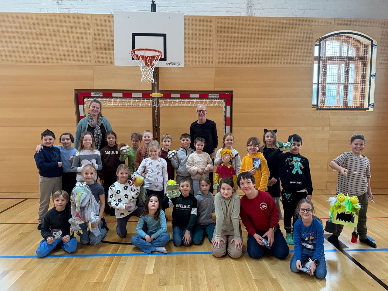 A group of children and adults pose for a photo in a gymnasium. The children are wearing costumes, and some are holding toys. The gymnasium has wooden flooring, and there is a basketball hoop and a goalpost.