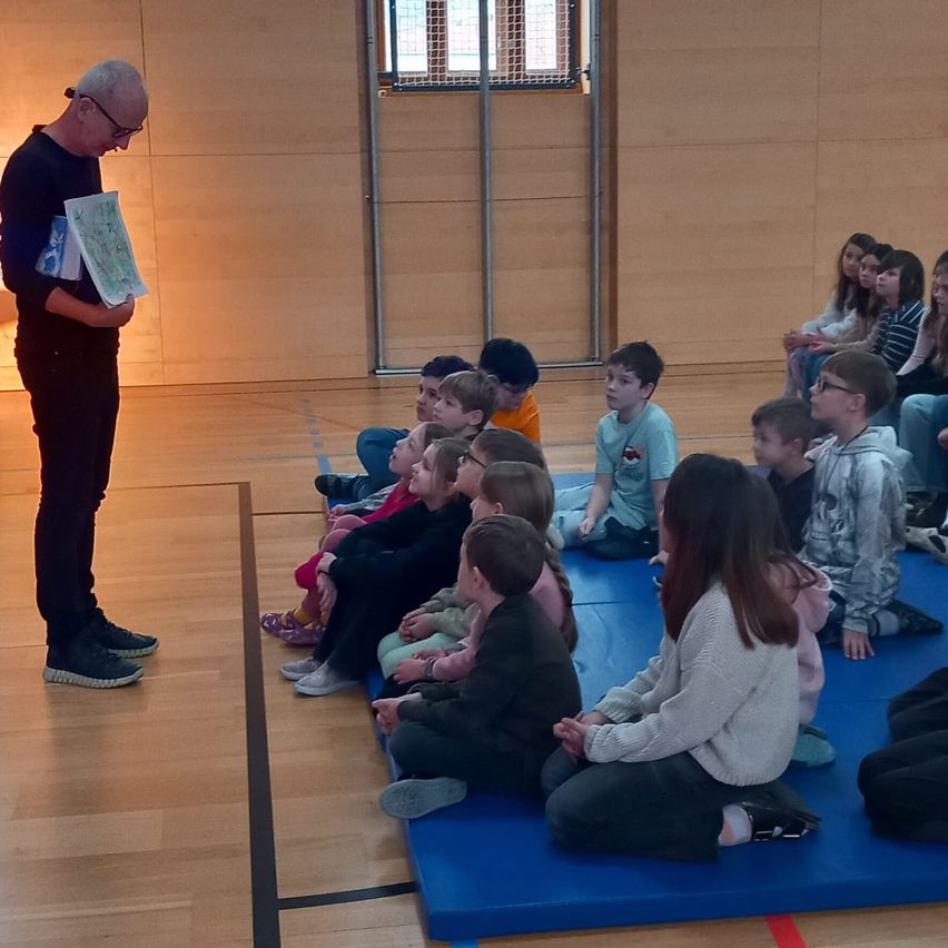 A man reads a book to a group of children sitting on a blue mat in a room with wooden floors and a window.
