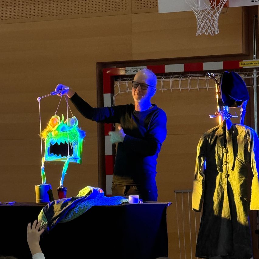 A person with glasses is performing a puppet show on a stage. The puppet is colorful with a monster-like appearance. The stage is lit with blue light.