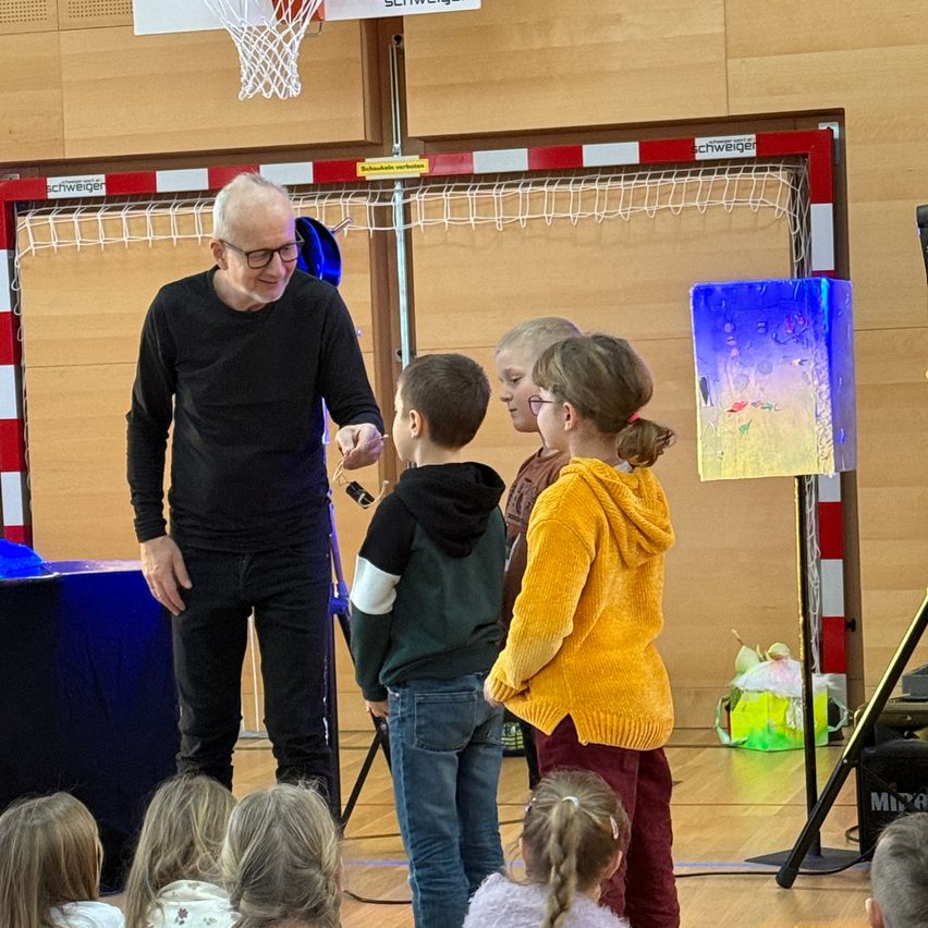 An older man in a black long-sleeve shirt is showing something to a group of children, who are listening intently. They are in a gymnasium with a basketball hoop in the background.