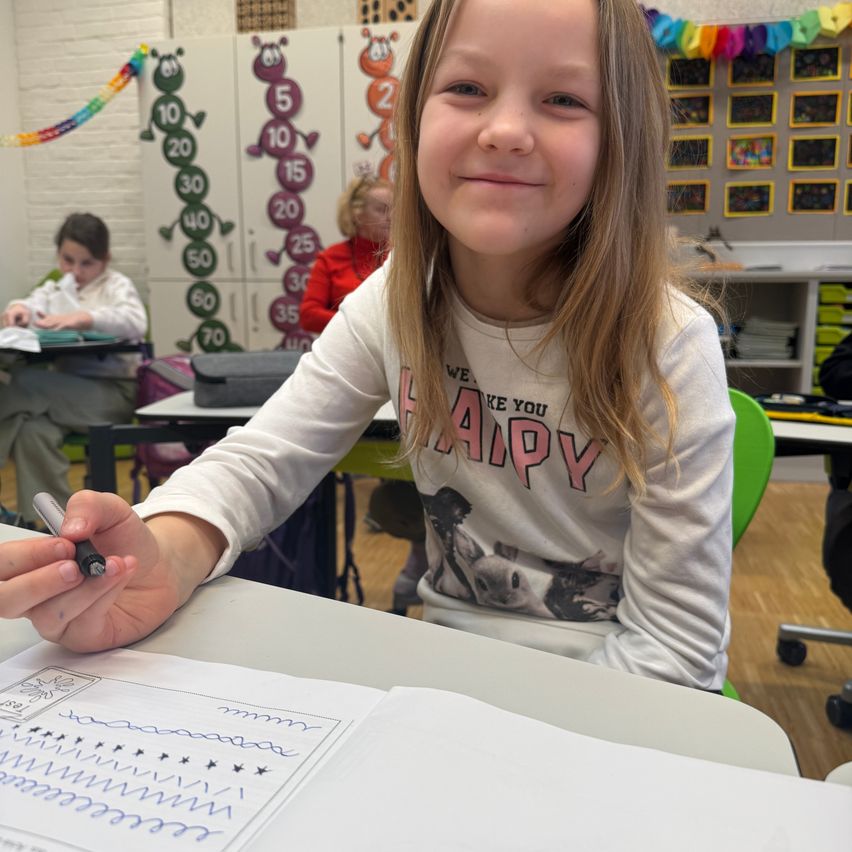 A young girl in a classroom is holding a pen and smiling while writing on a piece of paper. Another girl is sitting at a desk in the background.