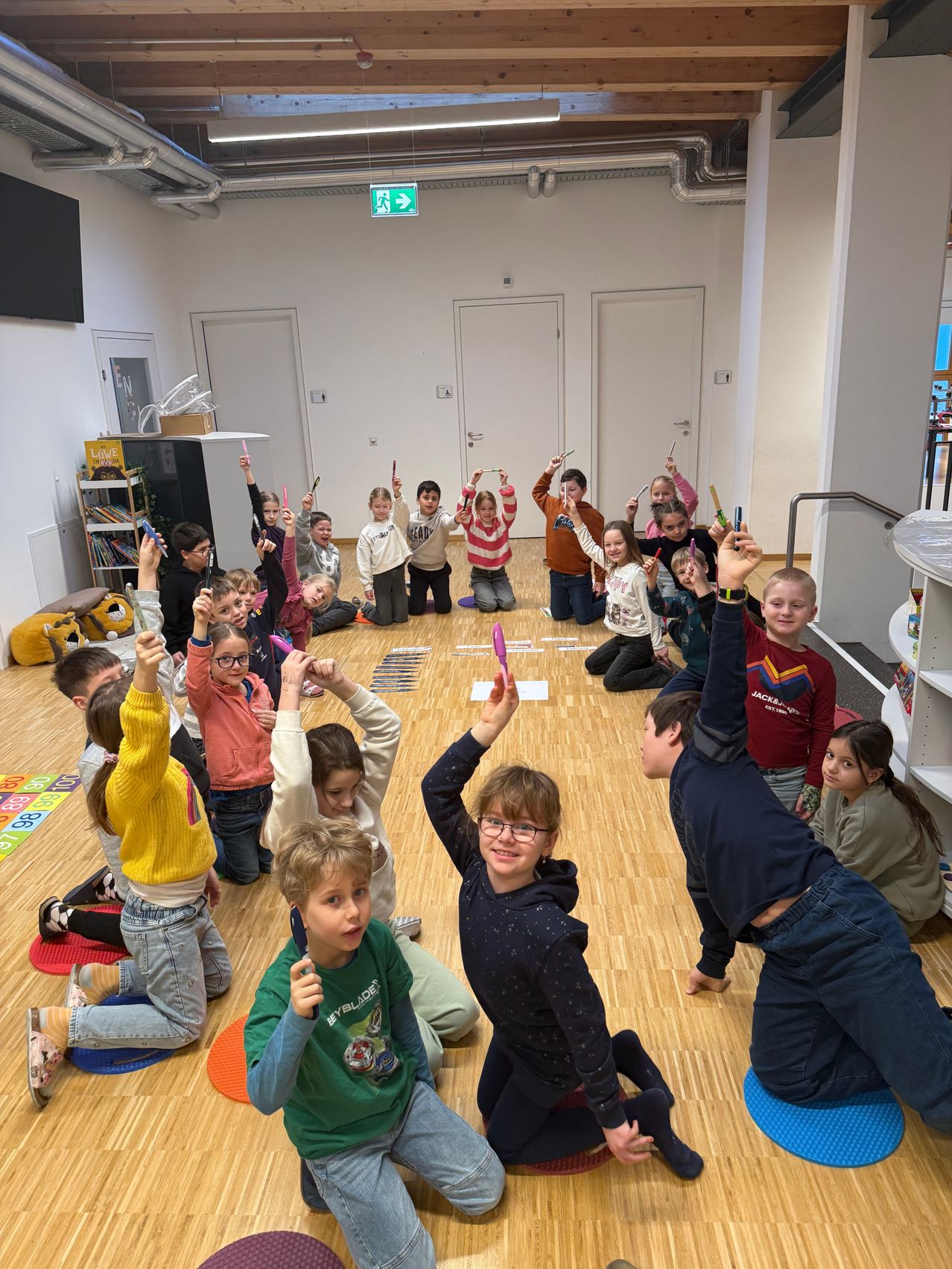 A group of children are kneeling and sitting on the floor, holding and playing with colorful objects. They are inside a room with white walls and wooden floors. Doors and shelves with books are visible.