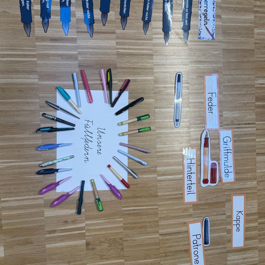 An array of pens and pencils with different names are displayed on a wooden surface. Each item is labeled with a name and has a distinct color.