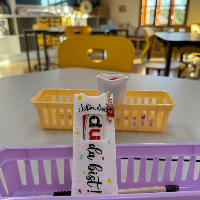 Ein Klassenzimmer mit einem Schreibtisch, auf dem ein violetter Korb mit einem Stiftetui mit der Aufschrift 'Big Listi' und einem kleinen Behälter mit einem roten Gegenstand steht. Dahinter stehen zwei gelbe Körbe auf einem weißen Tisch.