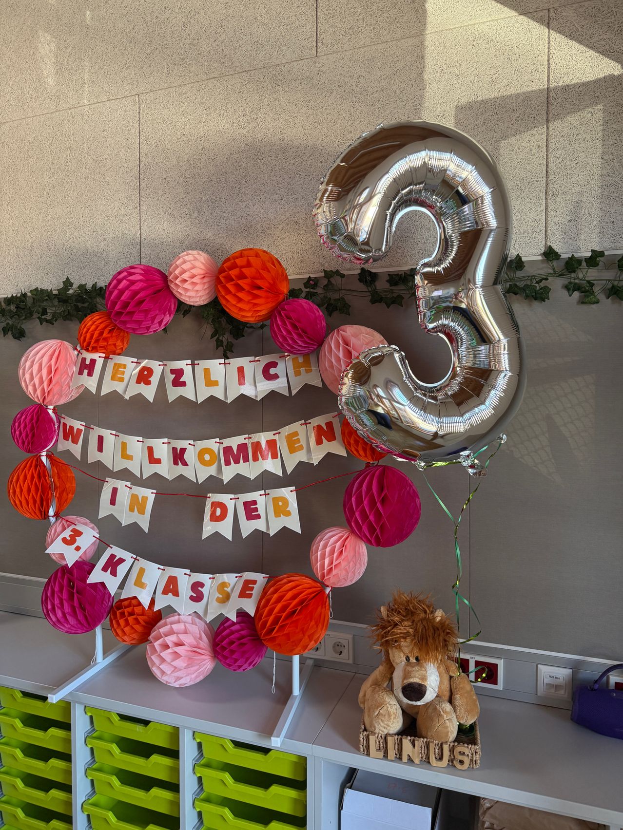 Eine bunte Ballondekoration mit einer silbernen Nummer drei, einem Banner mit der Aufschrift Herzlich Willkommen in der 3. Klasse und Papierkugeln in verschiedenen Farben. Unten steht ein brauner Plüschlöwe.